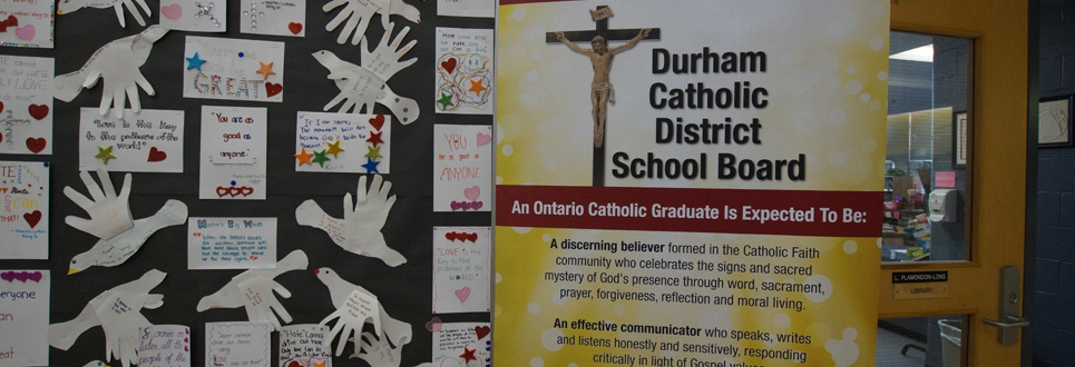 Stand up sign stating the Catholic Graduate Expectations beside a bulletin board with positive messages and doves made from traced hands.